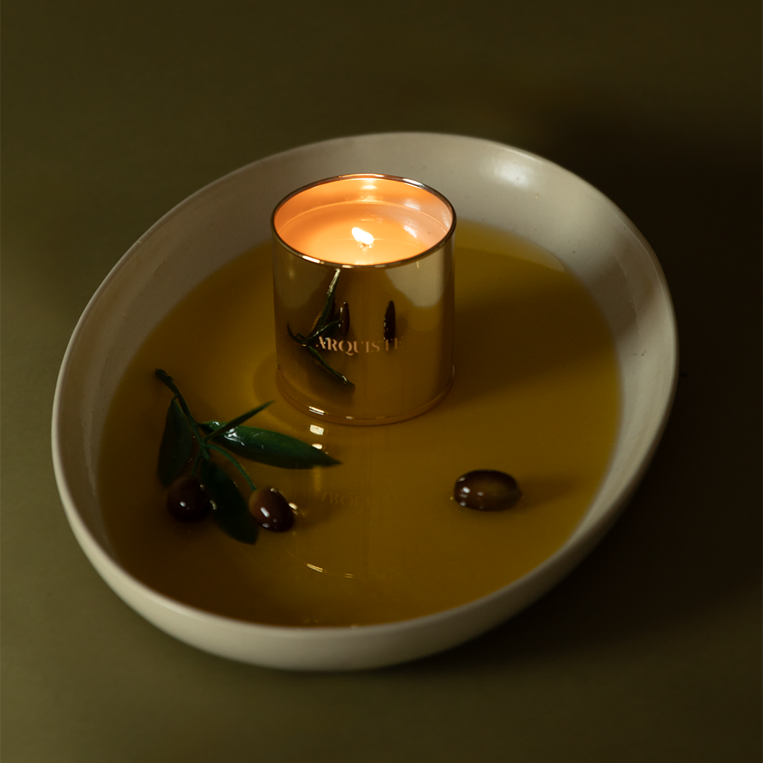 A lit metallic gold Arquiste candle in a white ceramic dish with olive branches and dark olives on a dark olive-green background.
