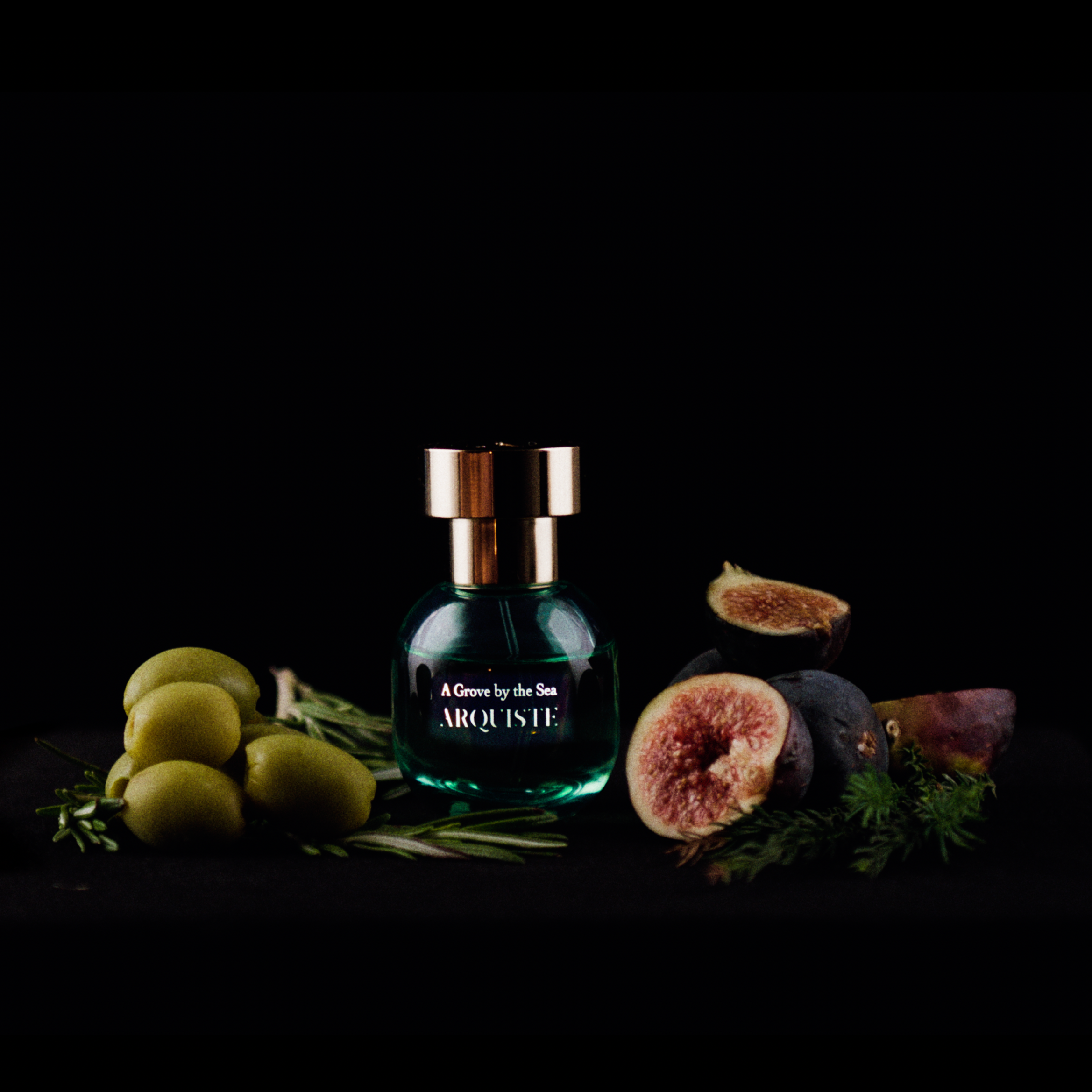A dark green glass perfume bottle with a gold cap and 'A GROVE BY THE SEA ARQUISTE' label, styled with fresh figs, olives, and rosemary on a black surface against a black background.