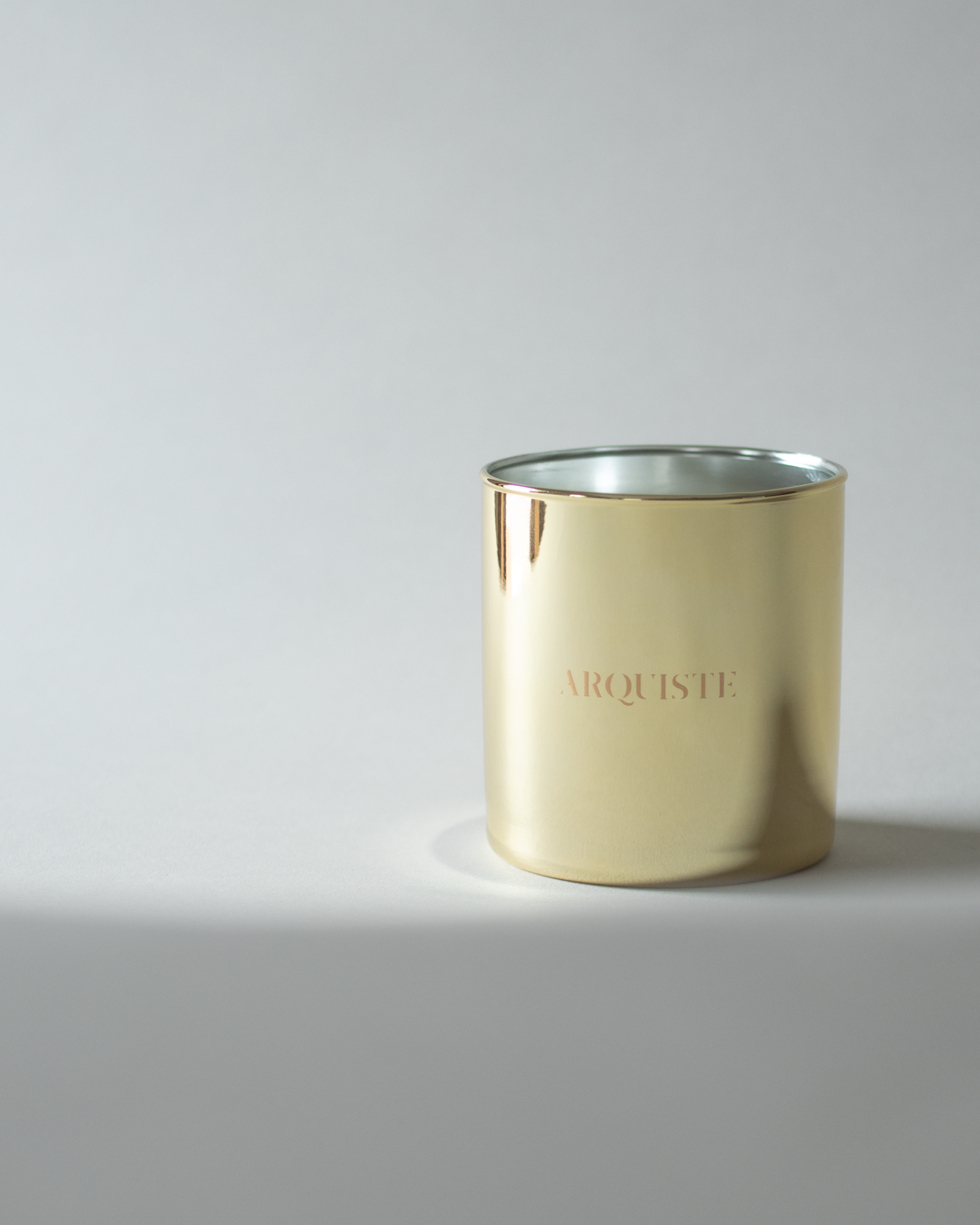 A metallic gold candle jar with the brand name 'ARQUISTE' printed in gold on the front, set against a neutral gray background.