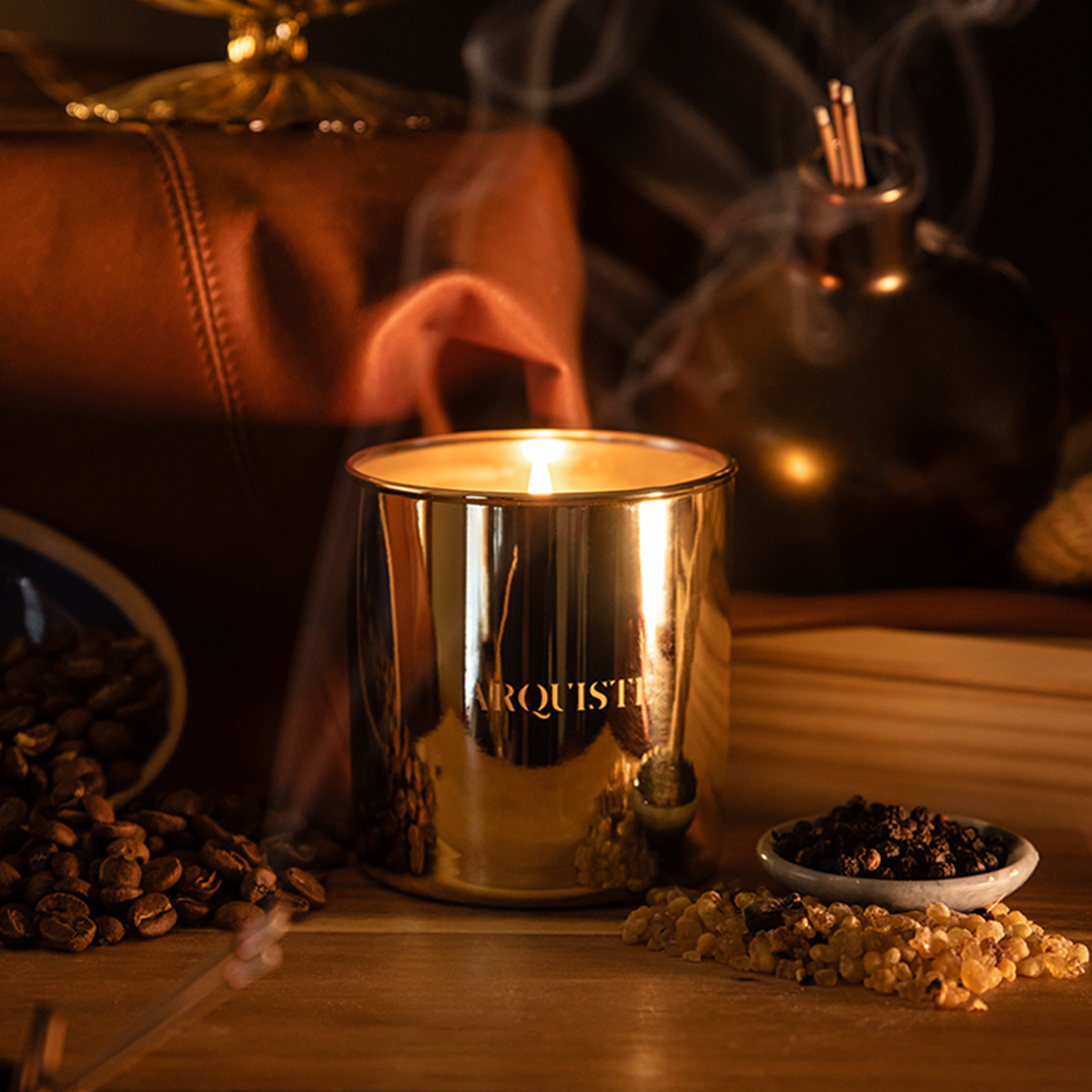 A metallic gold scented DARK GALLEON candle labeled 'ARQUISTE' sits lit on a wooden surface, surrounded by coffee beans, dried botanicals, and rising smoke in a warm, moody setting.