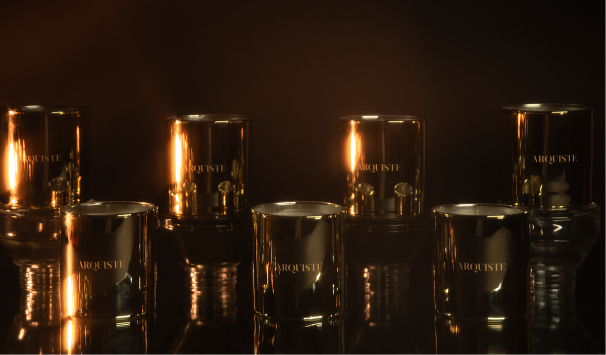 Six metallic gold glass candle jars with lit candles inside, arranged on a reflective dark surface against a black background. The brand name 'ARQUISTE' is visible on the front of the jars.