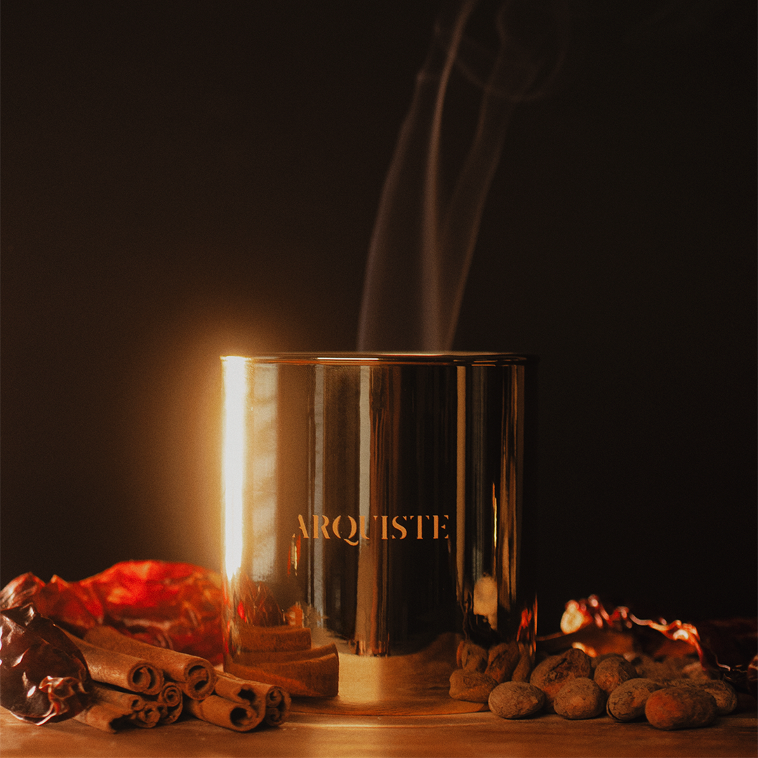 A metallic gold cylindrical candle holder or diffuser with the brand name 'ARQUISTE' printed in gold on the front, set on a wooden surface with warm glowing light, cinnamon sticks, dried flowers, and small nuts. Steam or smoke rises from the top against a dark black background.