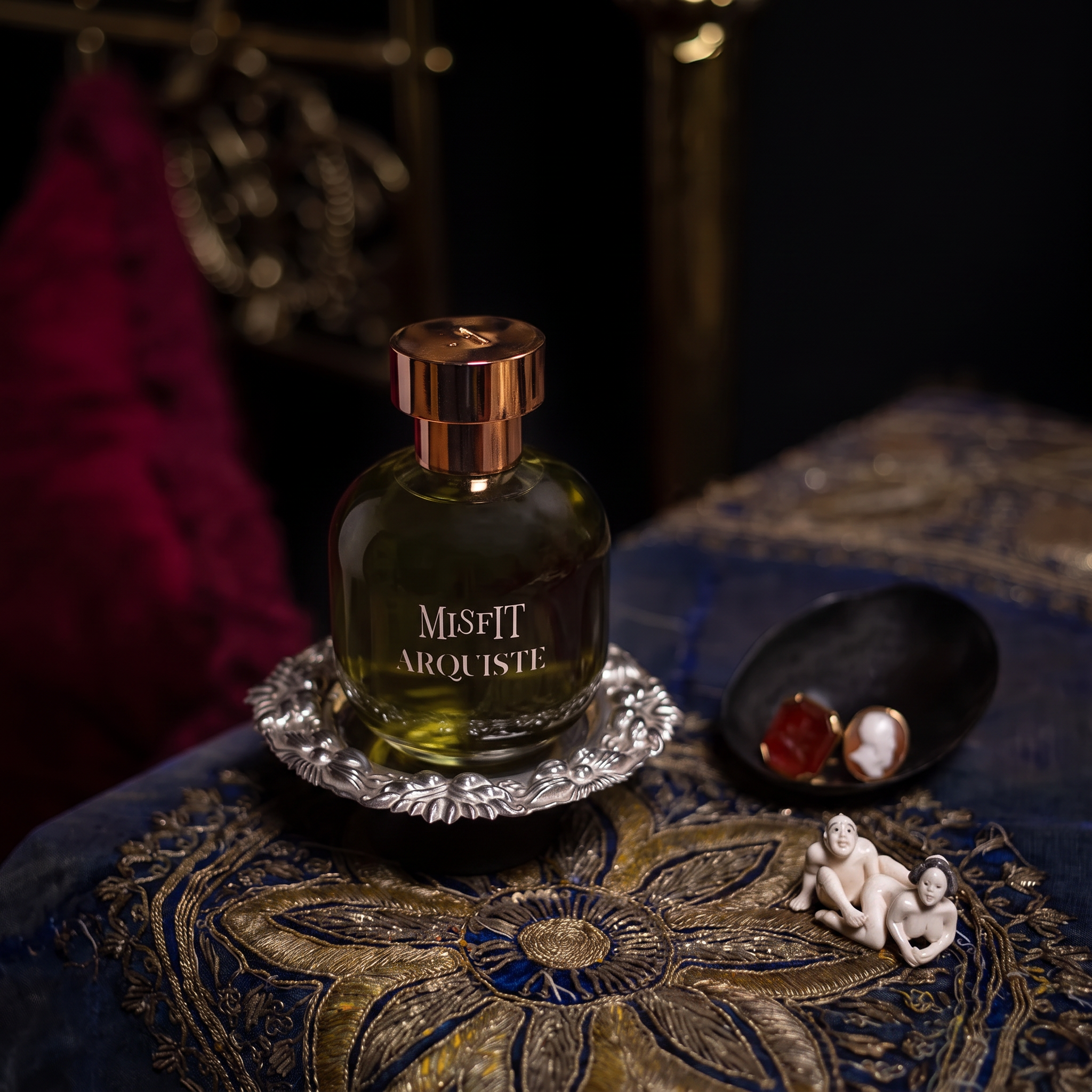 A bottle of Misfit Arquite perfume with a metallic rose gold cap and greenish-yellow liquid, displayed on a silver tray next to a small white netsuke figurine and a black dish with red and white antique rings, set on a blue and gold embroidered Kashmiri shawl.