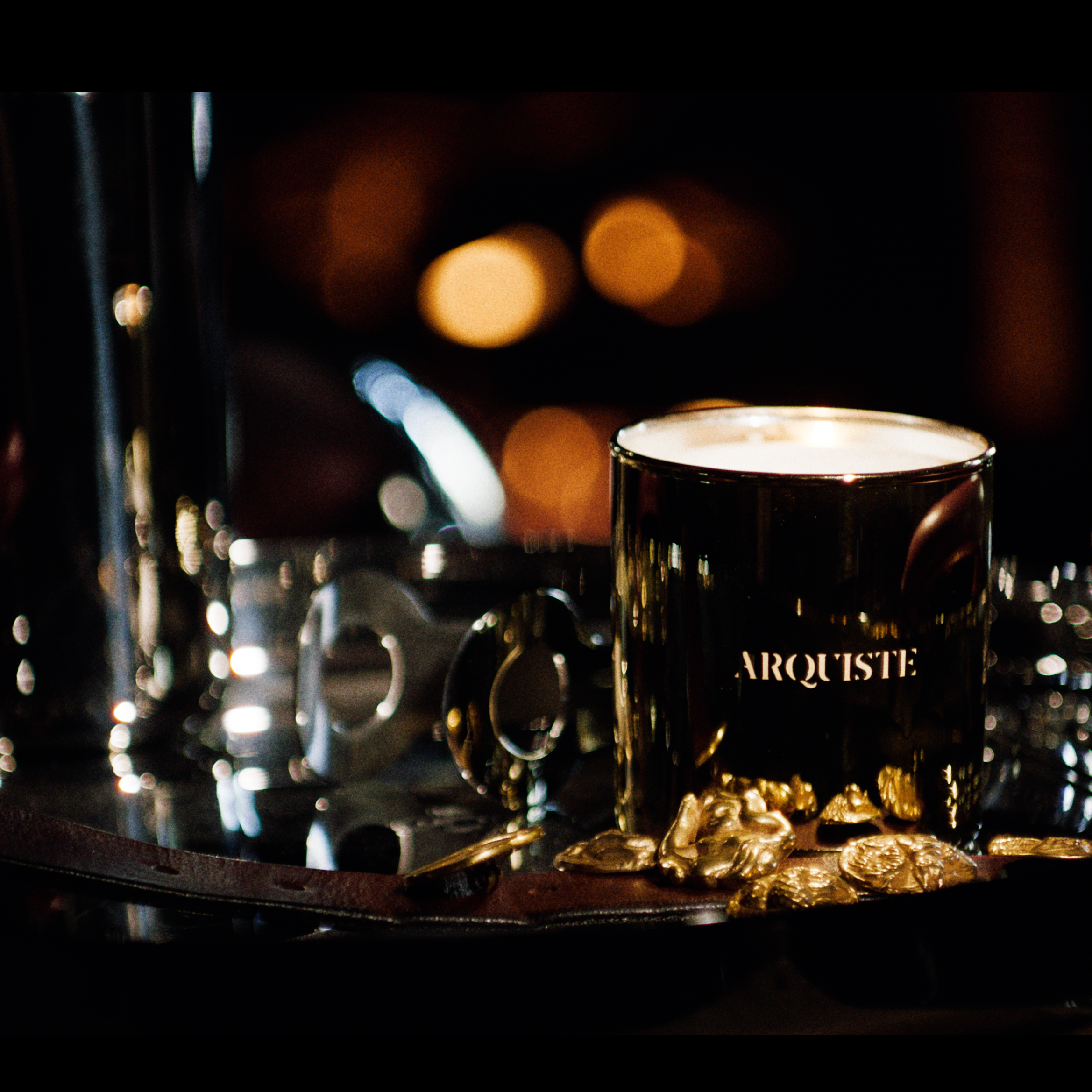 A gold glass jar candle with the brand name 'ARQUISTE' on the front, styled on a reflective dark tray with leather belts, gold coins and warm lights in the background.
