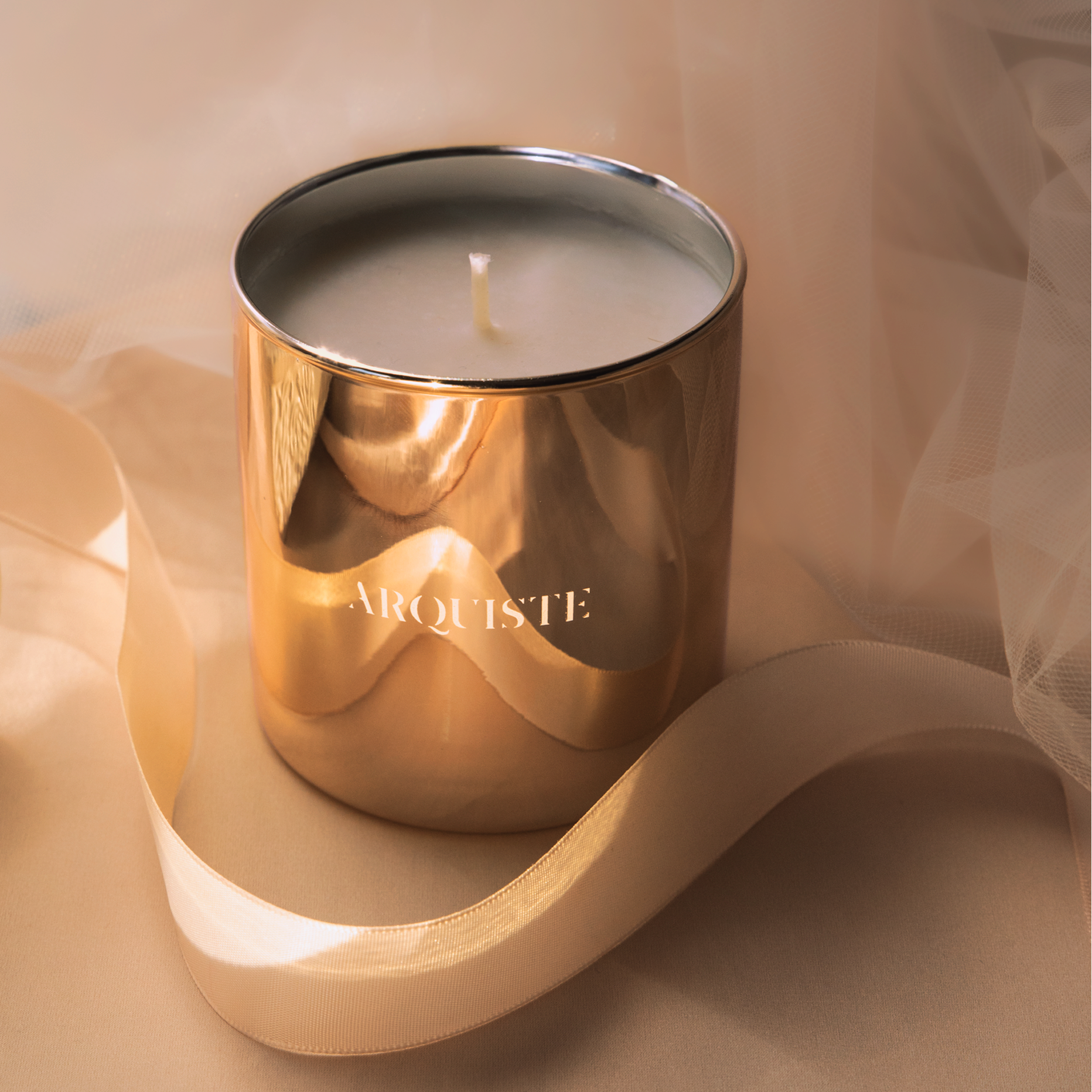 A metallic gold jar candle with the brand name 'ARQUISTE' printed in gold on the front, styled with a soft pink ribbon on a soft beige fabric background.