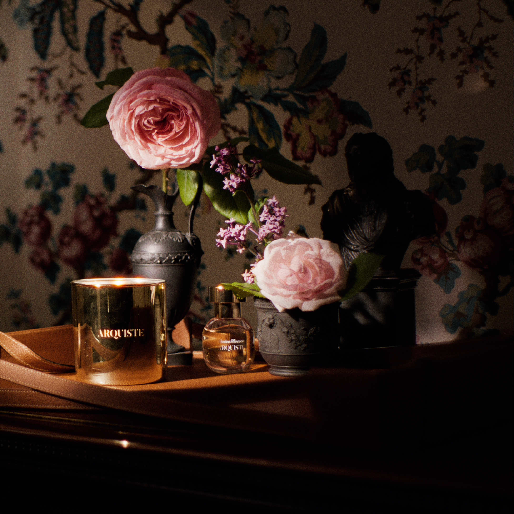 A styled interior scene with an antrique floral wallpaper, a dark wooden table, and a candlelit arrangement featuring a pink rose in a black basalt Wedgewood vase, an 18th century bust, a small pink flower in a pot, and a gold candle jar labeled 'ARQUISTE' on a leather tray.