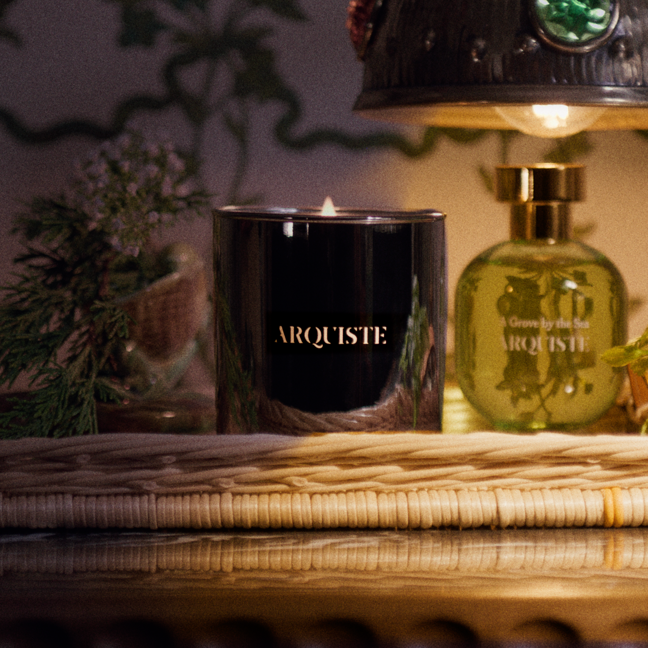 A styled tabletop scene with a gold candle labeled ARQUITSTE beside a green glass perfume bottle labeled A Grove by the Sea Arquiste, set in a warm dimly lit interior with a lamp, basket, and greenery.