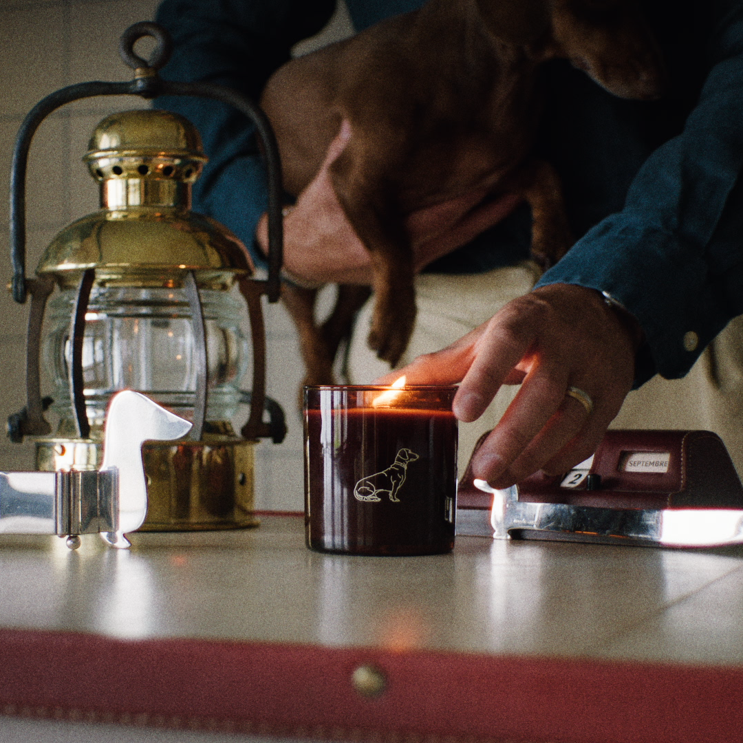 A lit candle in a amber glass jar sits on a tabletop next to a brass lantern and a small dachshund, being held by a person in a dim indoor setting.