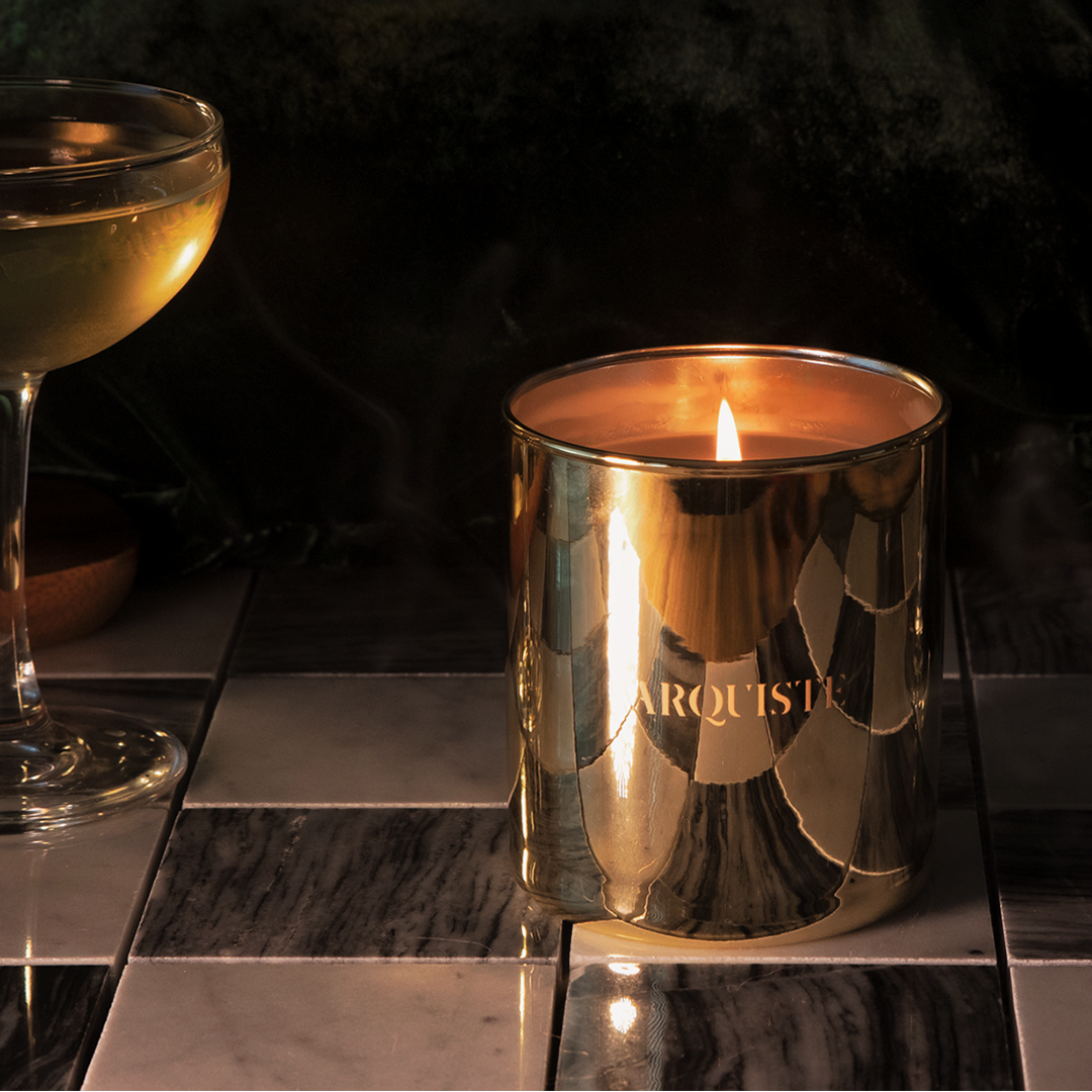 A metallic gold Arquiste candle in a reflective cylindrical holder, lit and placed on a dark tiled surface next to a coupe glass with pale liquid.