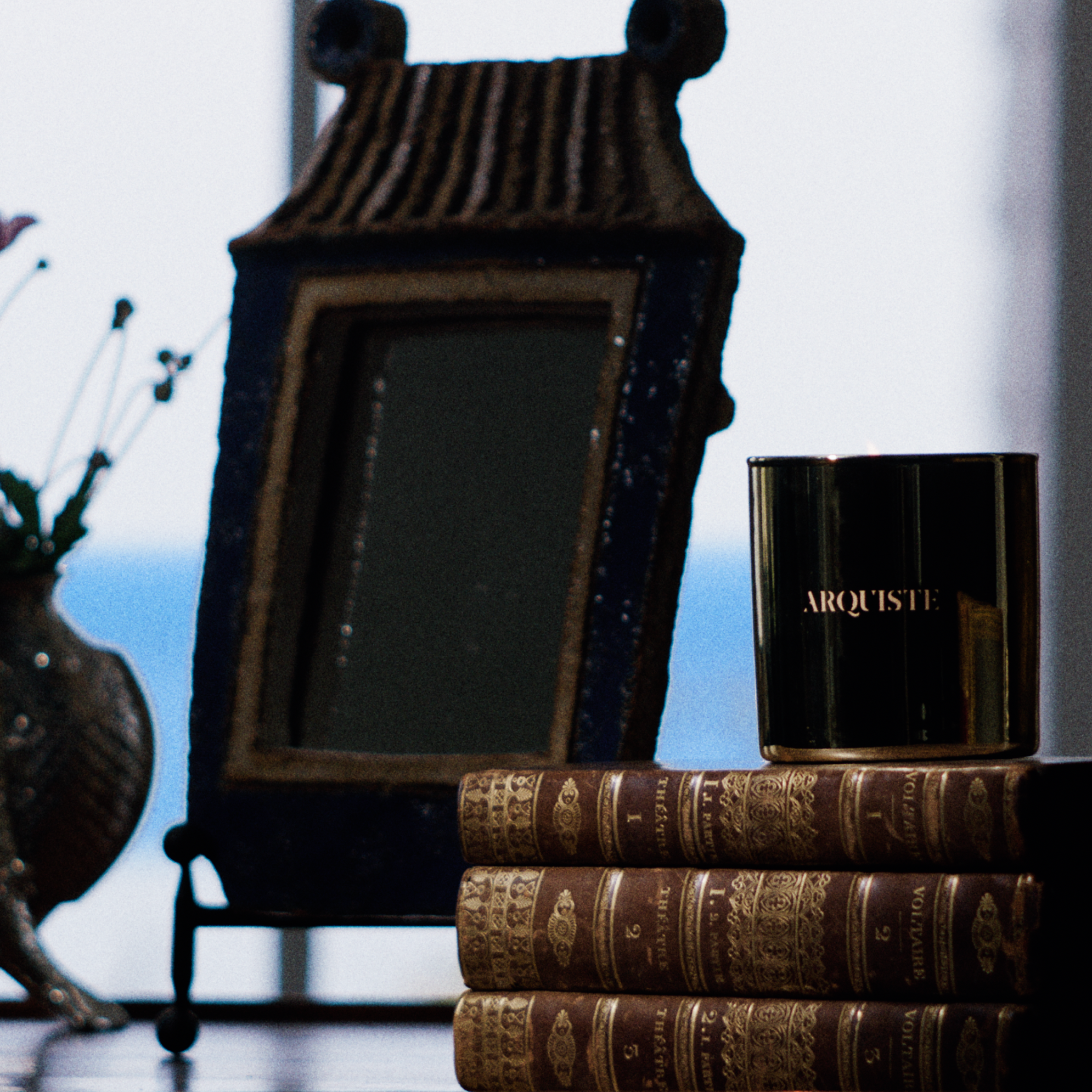 A dark glass candle jar with gold lettering reading 'ARQUISTE DARK GALLEON' sits on top of a stack of antique-style books next to a decorative vase with pink flowers and a Pagoda shaped picture frame, set against a bright window background.