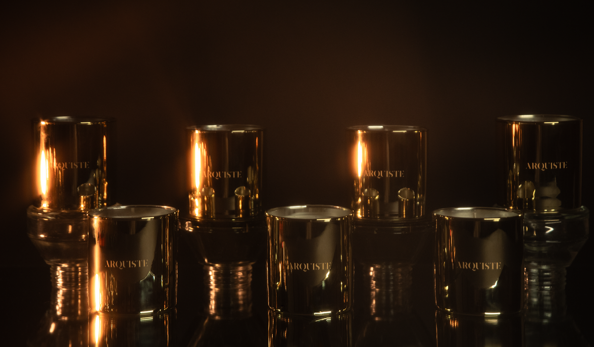 Six metallic gold glass candle jars with lit candles inside, arranged on a reflective dark surface against a black background. The brand name 'ARQUISTE' is visible on the front of the jars.