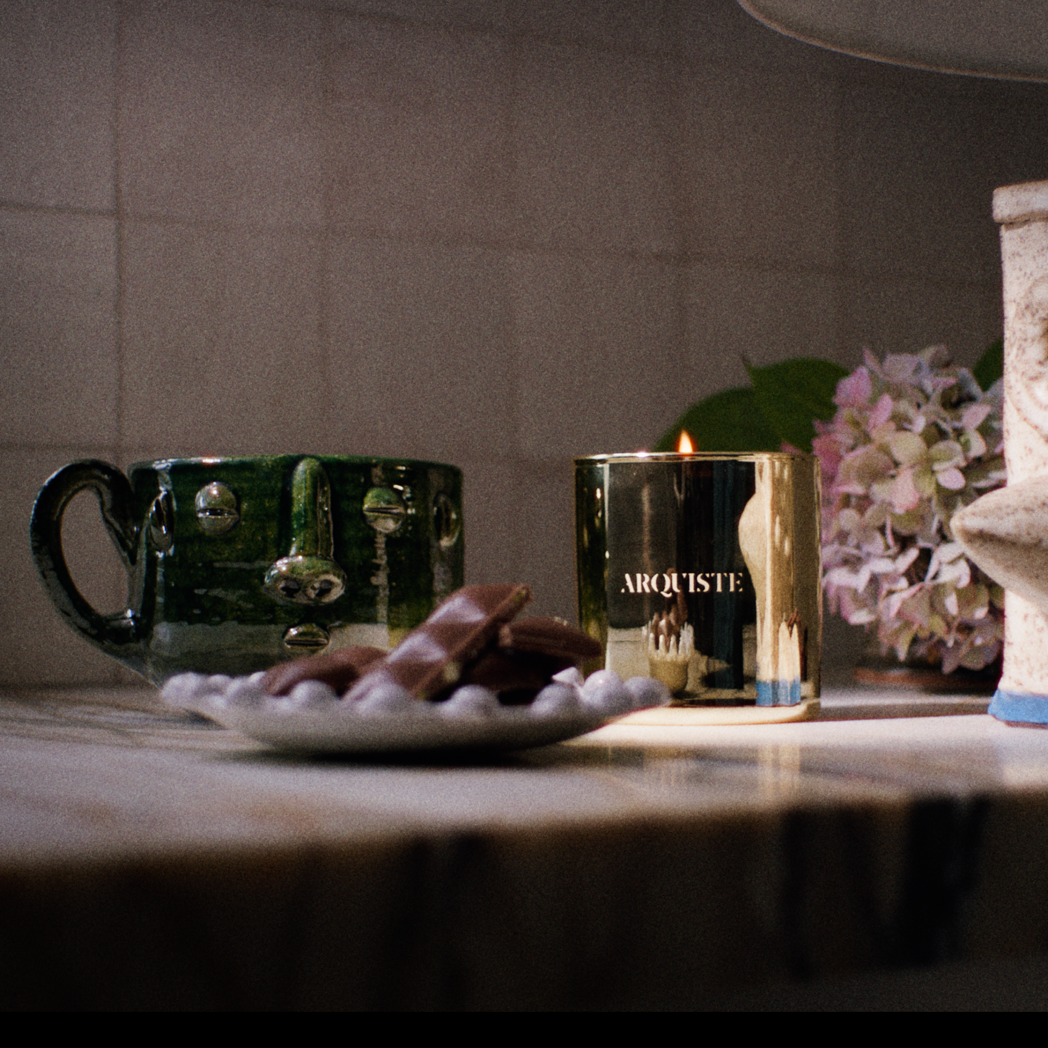 A lit gold candle jar with the brand name 'AIRQUISTE' visible on the front, styled on a marble countertop next to a green ceramic mug, a small plate of chocolates, and a ceramic bird figurine.