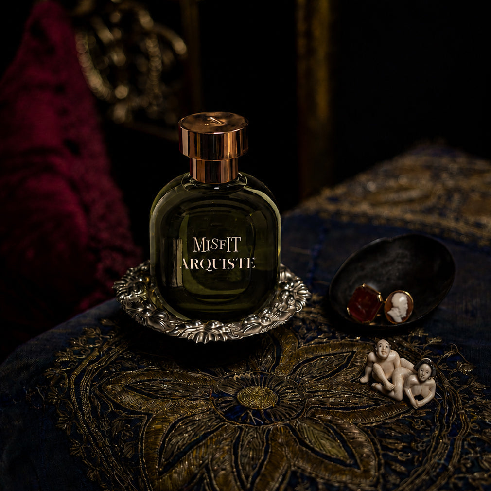 A dark green glass perfume bottle with a gold cap and 'MISFIT ARQUSITE' text, styled on a silver tray next to 19th century rings and a japanese netsuke figurine, on an ornate embroidered blue and gold textile from India.