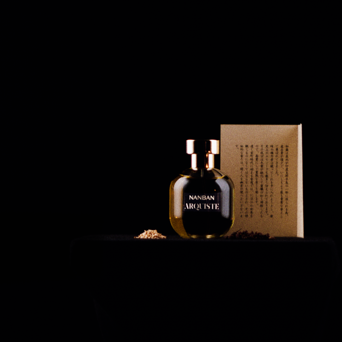 A dark amber ARQUISTE Nanban glass perfume bottle with gold-toned cap and lettering, standing beside a tan rectangular box  with sino-japanese script, on a black surface against a black background.