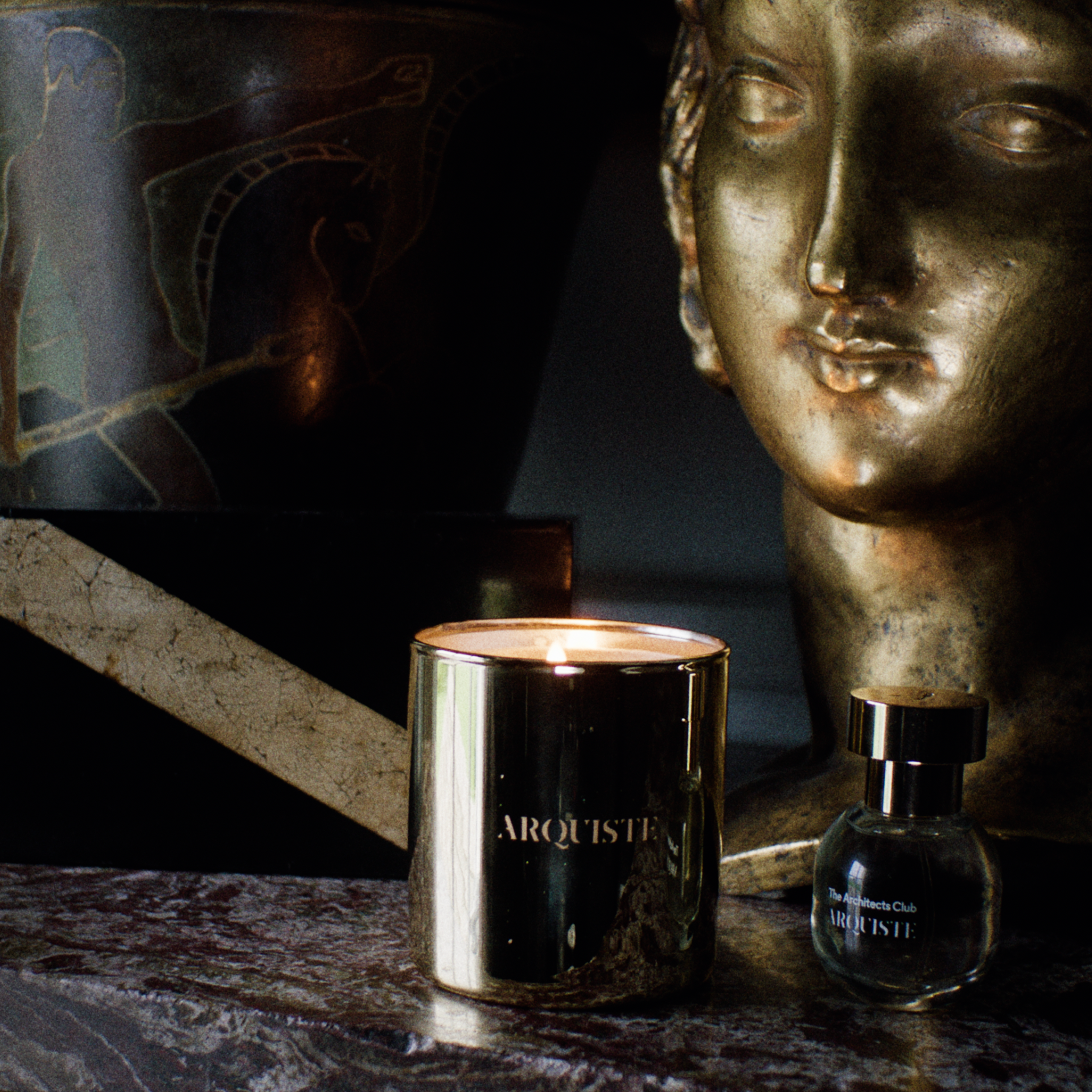 A lit metallic gold candle and a small dark glass perfume bottle with gold lettering, styled on a marble surface next to an Art Deco bust, vase and a black box in a moody interior setting.