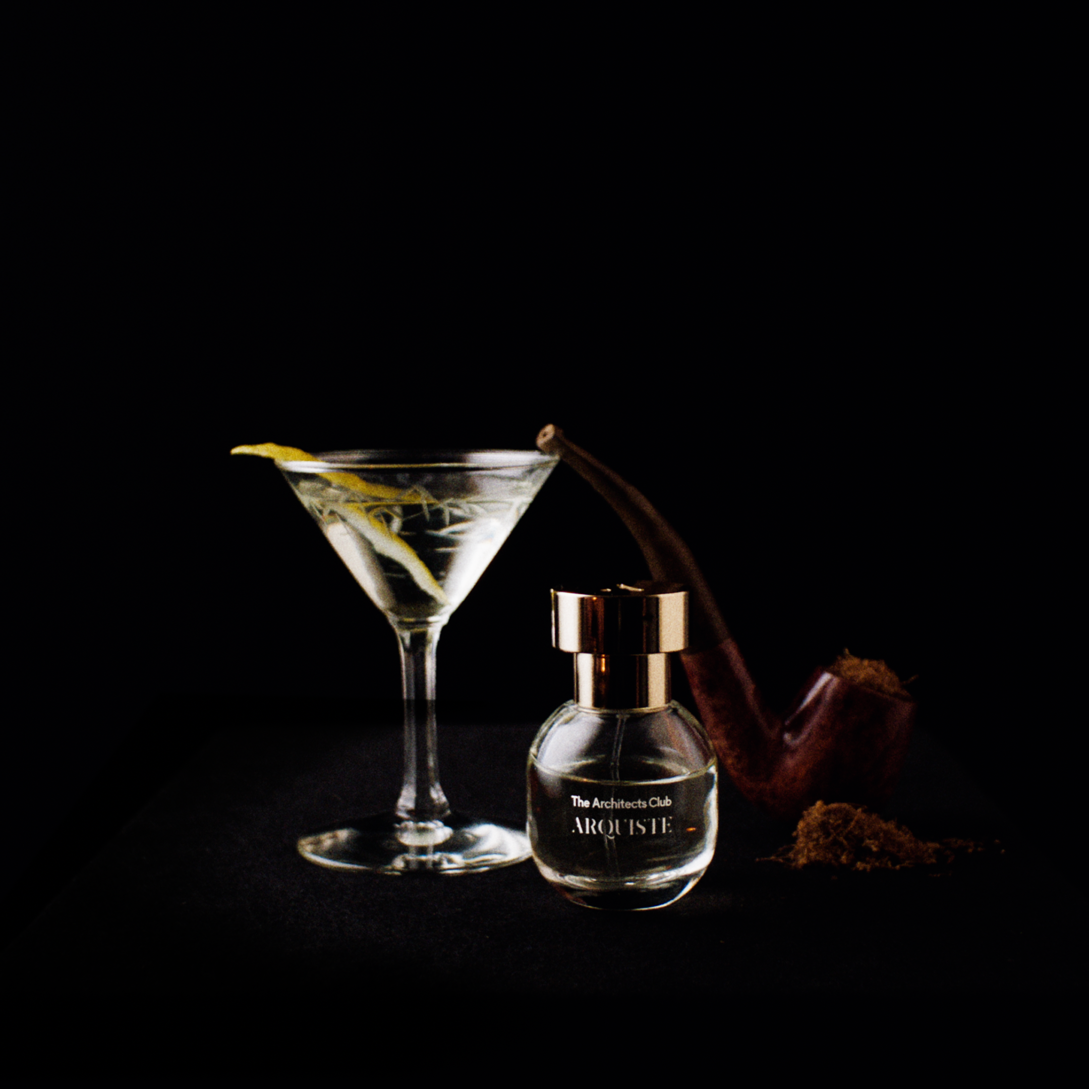 A small The Architects Club by Arquiste perfume bottle with a metallic gold cap sits on a black surface beside a martini glass with a lemon twist and a dark brown pipe, against a solid black background.