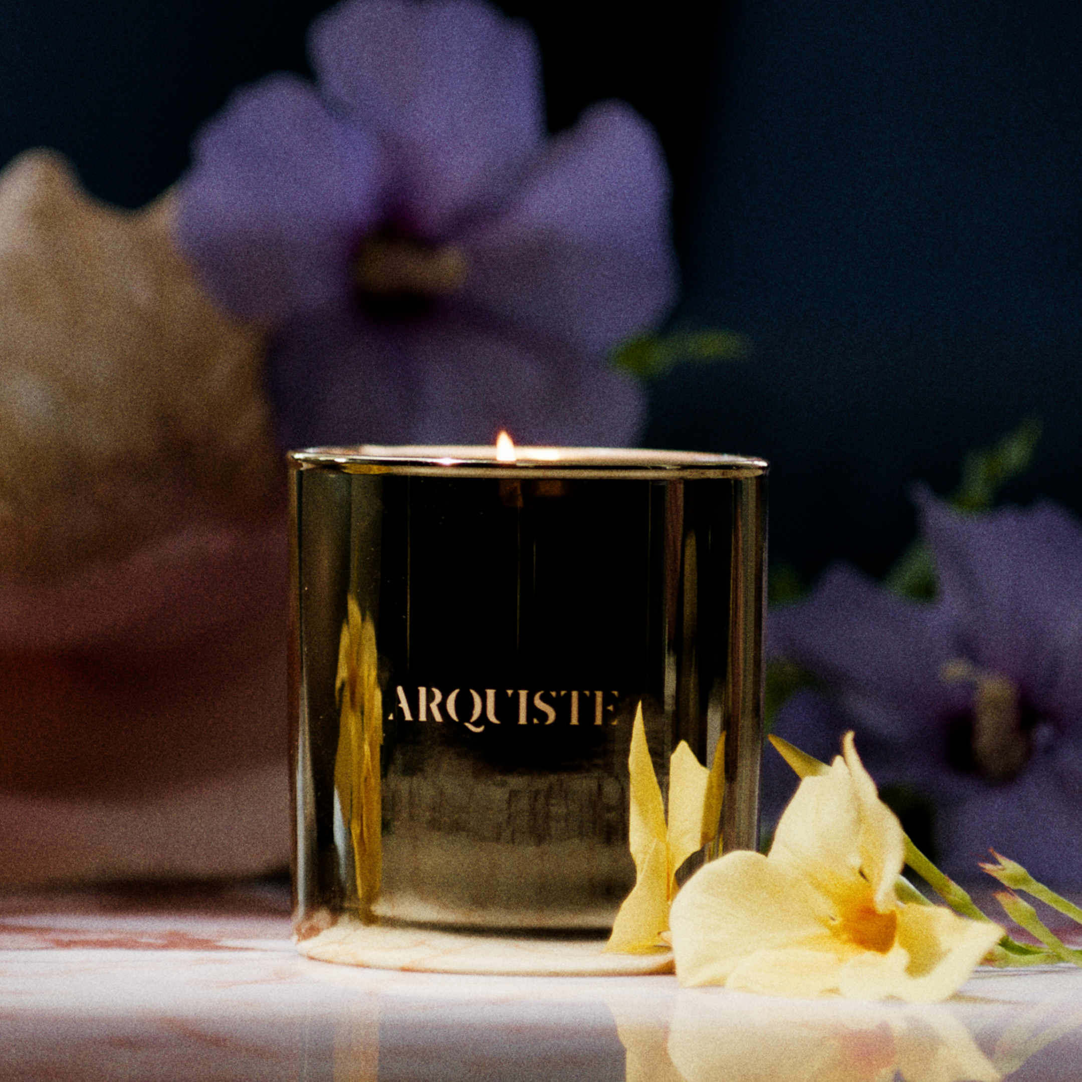 A gold metallic candle jar with the brand name 'ARQUISTE' printed in gold lettering, lit and set on a marble surface surrounded by yellow flowers, a seashell, and purple flowers in a dark, moody setting.
